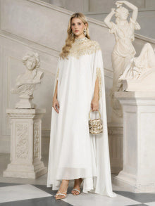 A woman in the Social Queen Beaded Tulle Caftan Cape Dress stands indoors between classical statues, holding a small handbag and wearing heeled sandals—a sophisticated choice for an evening event.