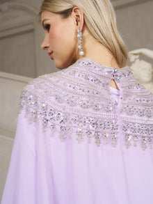 A woman with long blonde hair wears the Social Queen Beaded Tulle Caftan Cape Dress adorned with lavender beads, silver sequins, and a long pearl earring.