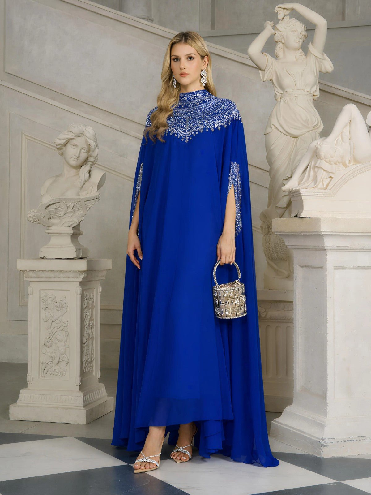 A woman in a Social Queen Beaded Tulle Caftan Cape Dress stands indoors holding a small ornate handbag, surrounded by classical marble statues and art decor.