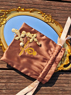 The Bezel-Set Crystal Cross Earrings by Social Queen, elegant wedding guest jewelry, rest on a brown satin pouch with gold embroidery, displayed on a gold-trimmed mirror atop a wooden surface.