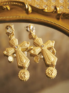 Two Bezel-Set Crystal Cross Earrings by Social Queen, featuring intricate patterns and round medallions with small pearls—ideal for wedding guests—are displayed on a reflective gold-framed surface.
