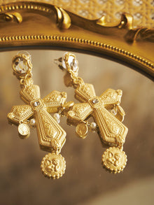 Two Bezel-Set Crystal Cross Earrings by Social Queen, featuring intricate patterns and round medallions with small pearls—ideal for wedding guests—are displayed on a reflective gold-framed surface.