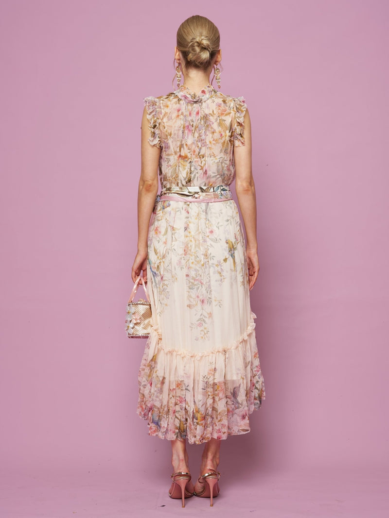 A woman faces away, wearing the Bird Floral Print Silk Dress by Social Queen. She pairs it with pink heels and holds a small purse against a pink background, her hair styled in a bun.