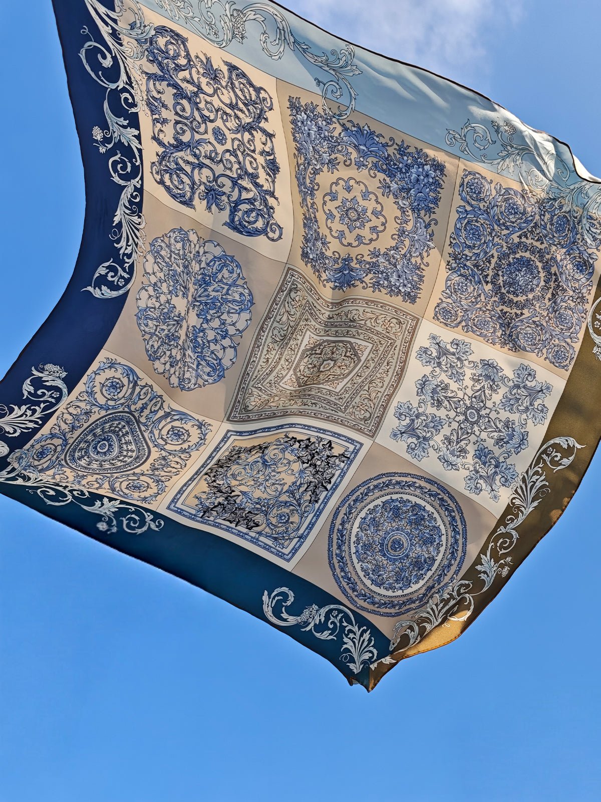 The Social Queen Bohemian Print Silk Scarf, featuring blue and beige intricate designs, is displayed suspended against a clear blue sky.