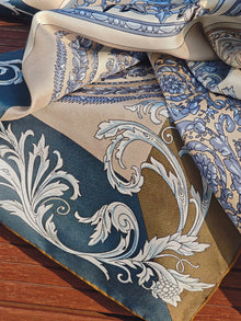 A close-up of the Social Queen Bohemian Print Silk Scarf features intricate blue, beige, and gold floral and ornamental patterns, displayed on a wooden surface.