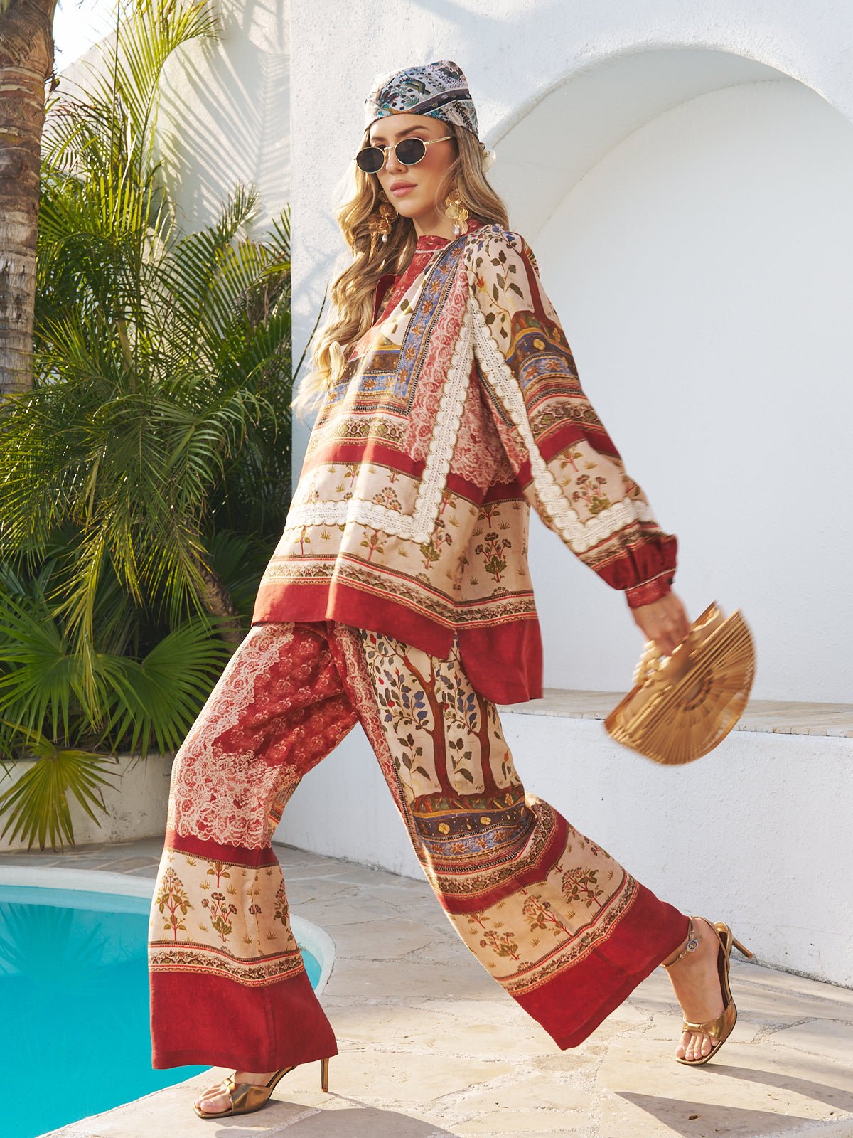 A woman walks by a pool wearing sunglasses, heeled sandals, and the Social Queen Bohemian Print Silk Scarf, paired with loose-fitting clothes and a bamboo handbag. Lush plants and white walls are in the background.