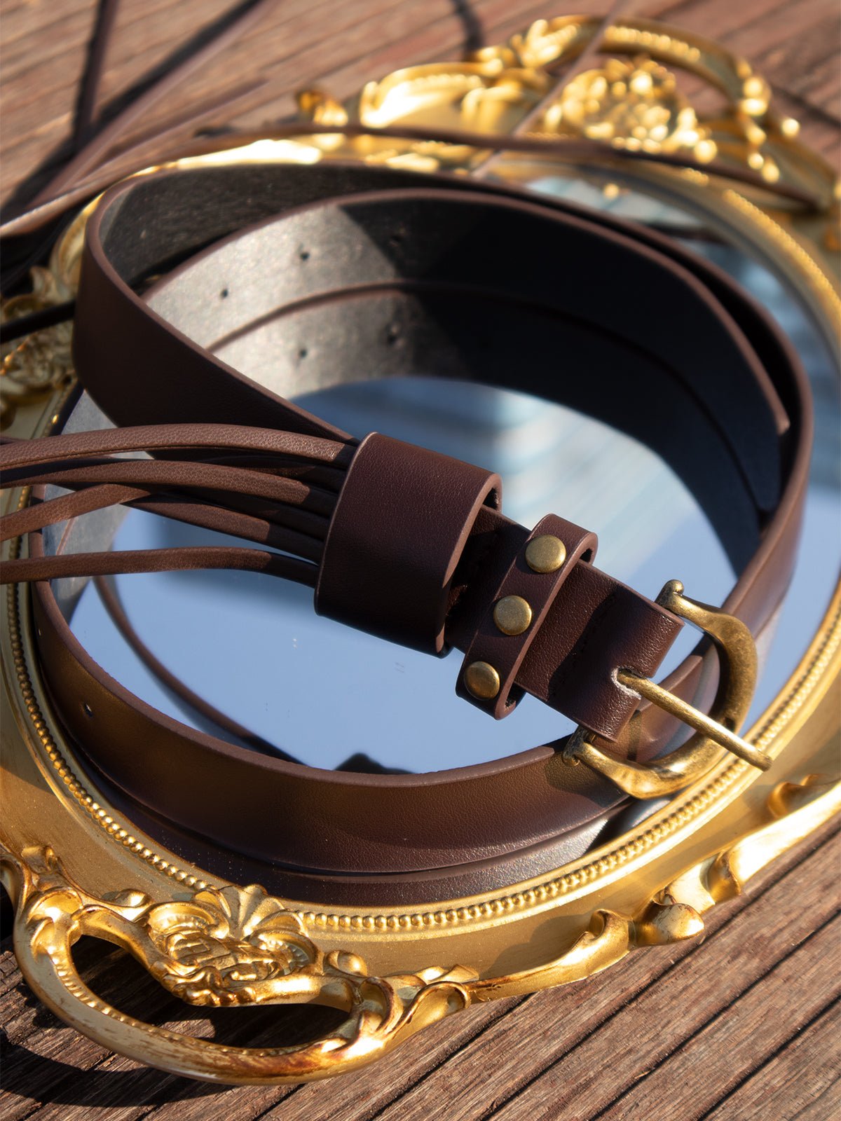 The Bohemian Tassel Leather Belt with D-Ring by Social Queen, featuring brown leather and brass accents, is coiled atop an ornate gold-framed mirror for a stylish Bohemian touch on the wooden surface.