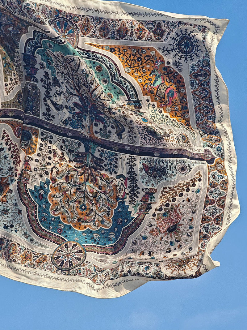 The Boho Silk Twill Scarf by Social Queen features vibrant floral and geometric patterns with delicate embroidery, fluttering gracefully against a clear blue sky.