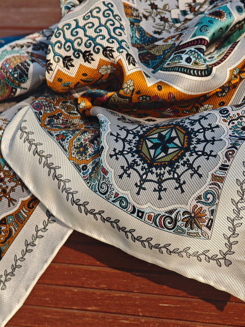 The Social Queen Boho Silk Twill Scarf, featuring intricate blue, yellow, and white geometric and floral prints, is draped over a wooden surface.