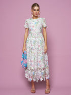 A woman poses against a pink background in Social Queen’s Botanical Embroidered Cutout Dress, featuring an A-line silhouette and short sleeves. She accessorizes with a colorful textured handbag, sandals, and statement earrings.