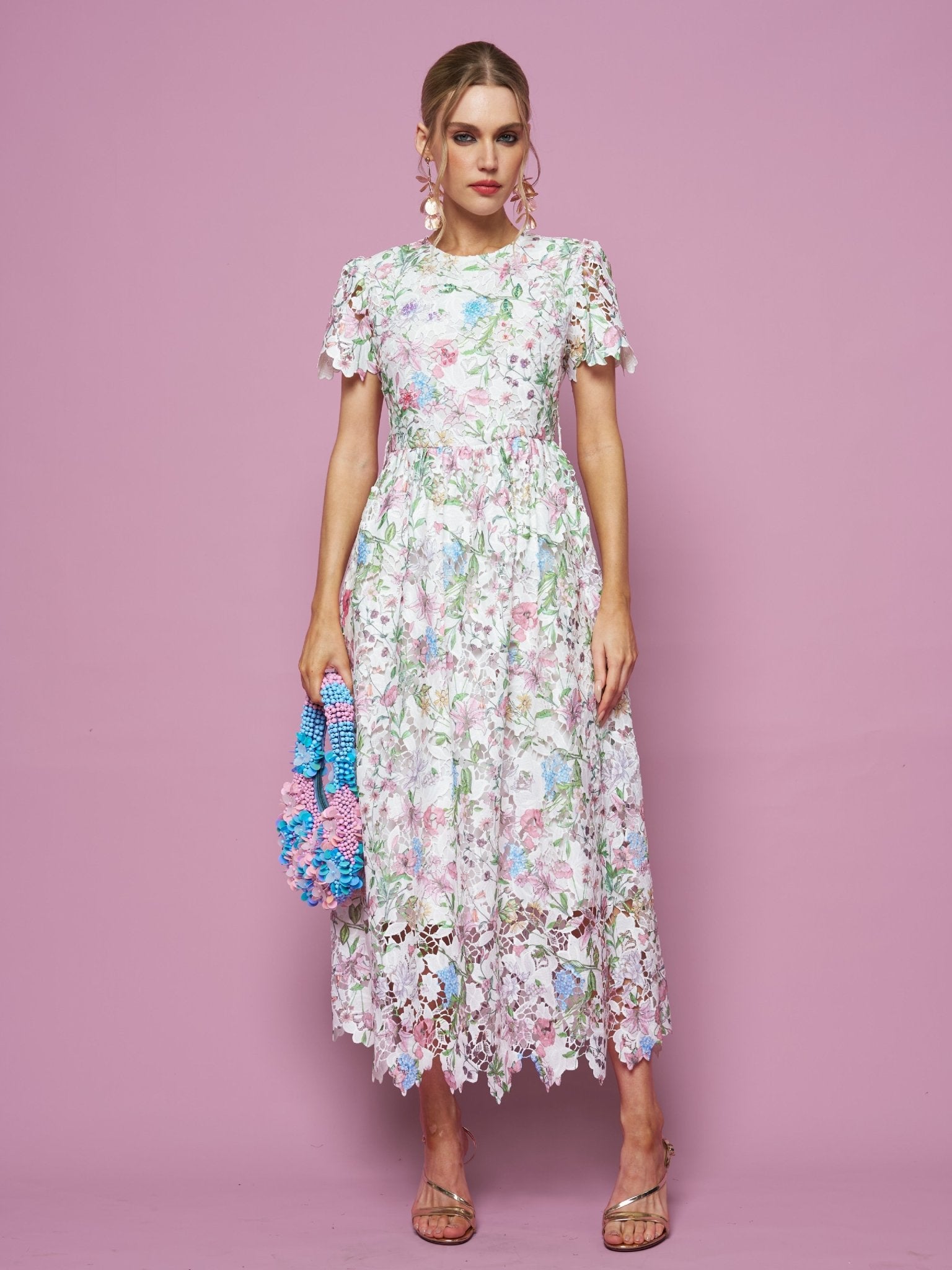 A woman poses against a pink background in Social Queen’s Botanical Embroidered Cutout Dress, featuring an A-line silhouette and short sleeves. She accessorizes with a colorful textured handbag, sandals, and statement earrings.