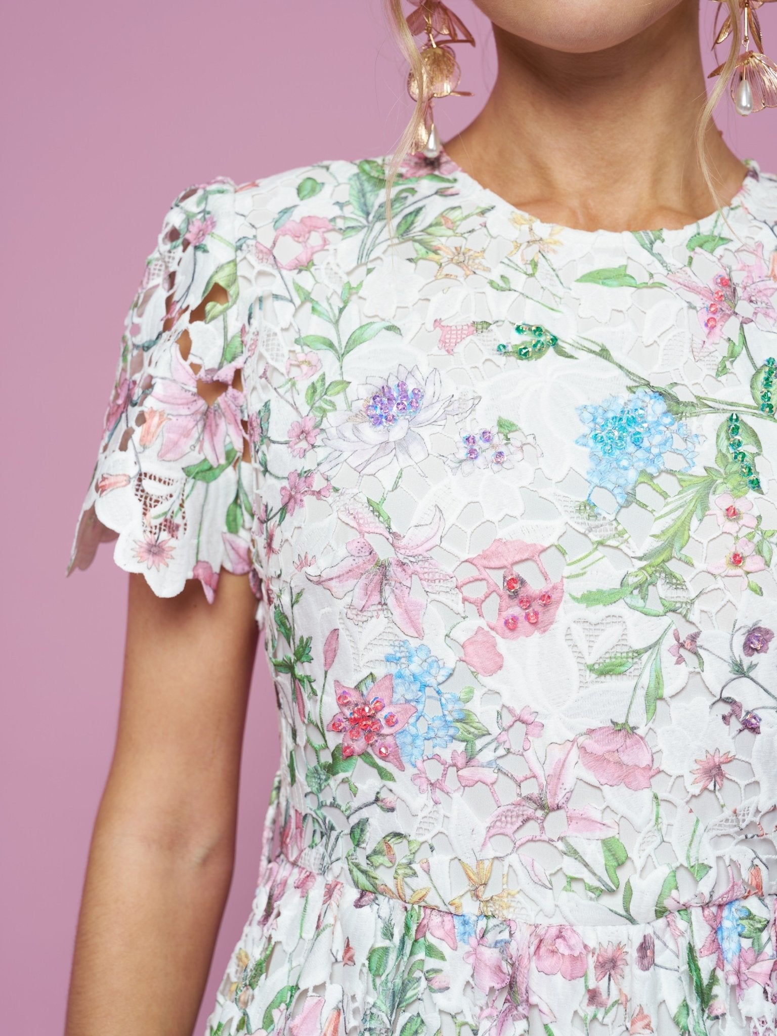 A woman wears the Social Queen Botanical Embroidered Cutout Dress, featuring white lace and floral embroidery with scalloped sleeves and an A-line silhouette, paired with large gold earrings against a pink background.