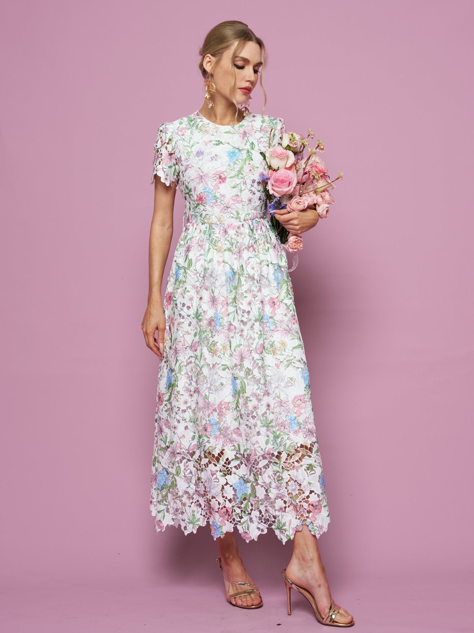 Woman wearing the Social Queen Botanical Embroidered Cutout Dress, featuring an A-line silhouette, holds a bouquet of pink flowers against a pink backdrop, paired with gold high-heeled sandals.