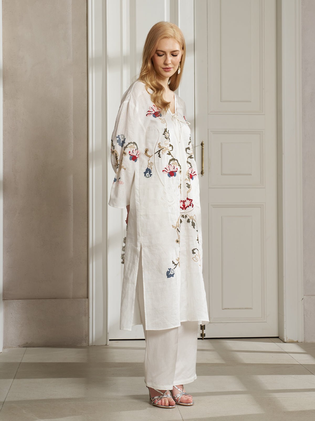 A woman models the Social Queen Botanical Embroidered Linen Top with white pants and pink sandals, standing sideways indoors by a closed white door, highlighting its ethnic-inspired embroidery and resort style.