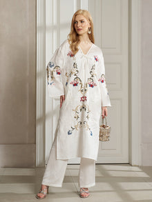 A woman stands indoors wearing the Social Queen Botanical Embroidered Linen Top, styled with white pants, silver sandals, and a woven handbag for an elegant resort-inspired look.
