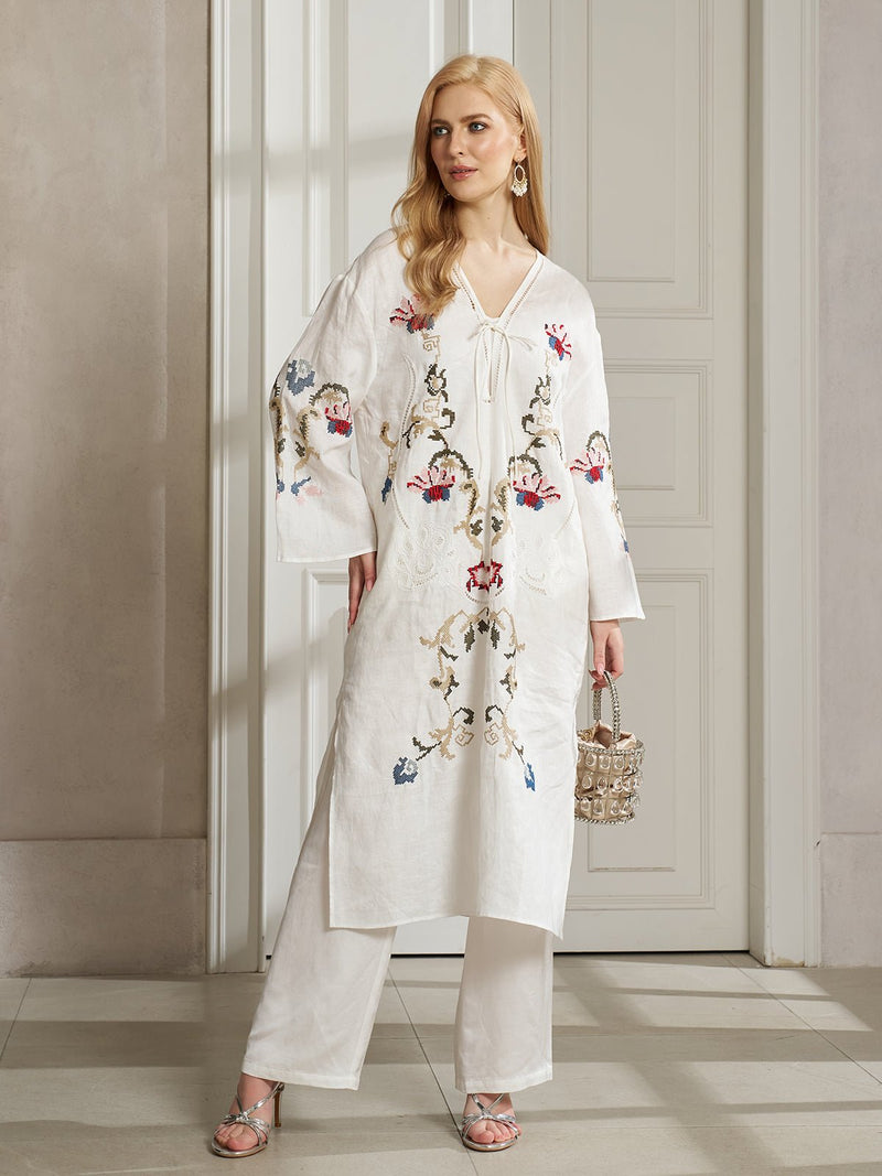 A woman stands indoors wearing the Social Queen Botanical Embroidered Linen Top, styled with white pants, silver sandals, and a woven handbag for an elegant resort-inspired look.