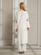 A woman with long blonde hair, wearing the Social Queen Botanical Embroidered Linen Top, stands indoors facing away in a resort-style white outfit, holding a small metallic handbag.