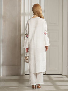 A woman with long blonde hair, wearing the Social Queen Botanical Embroidered Linen Top, stands indoors facing away in a resort-style white outfit, holding a small metallic handbag.