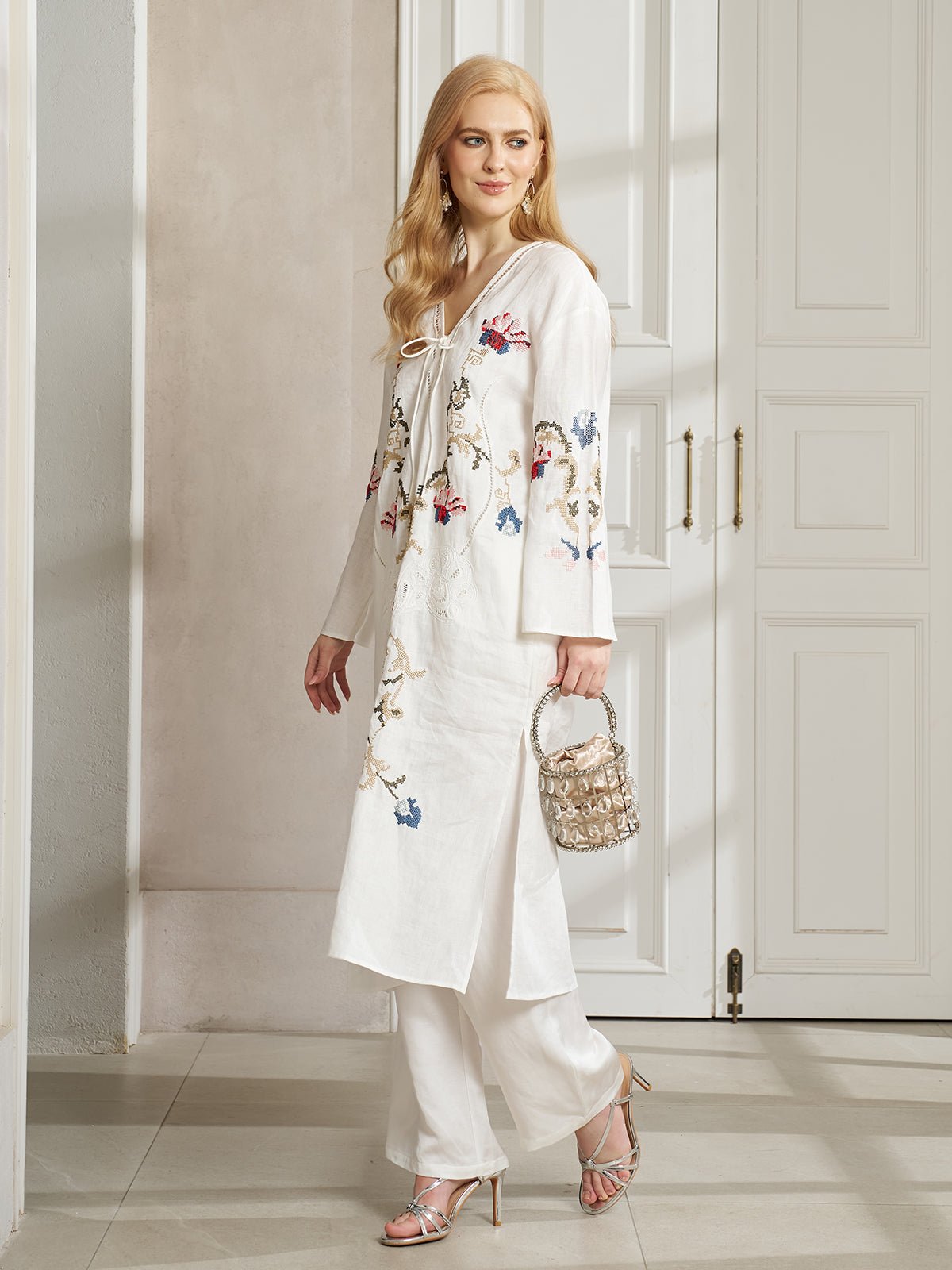 A woman stands indoors wearing Social Queen's Botanical Embroidered Linen Top and pants, paired with silver heels and a small handbag, showcasing elegant ethnic-inspired embroidery.
