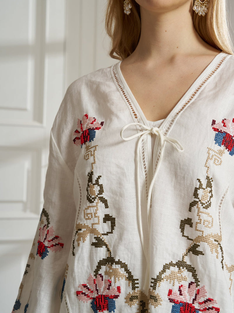 A woman models the Social Queen Botanical Embroidered Linen Top, featuring a V-neck tie and red, blue, and beige embroidery. The cropped photo emphasizes the resort-style appeal of this chic blouse.