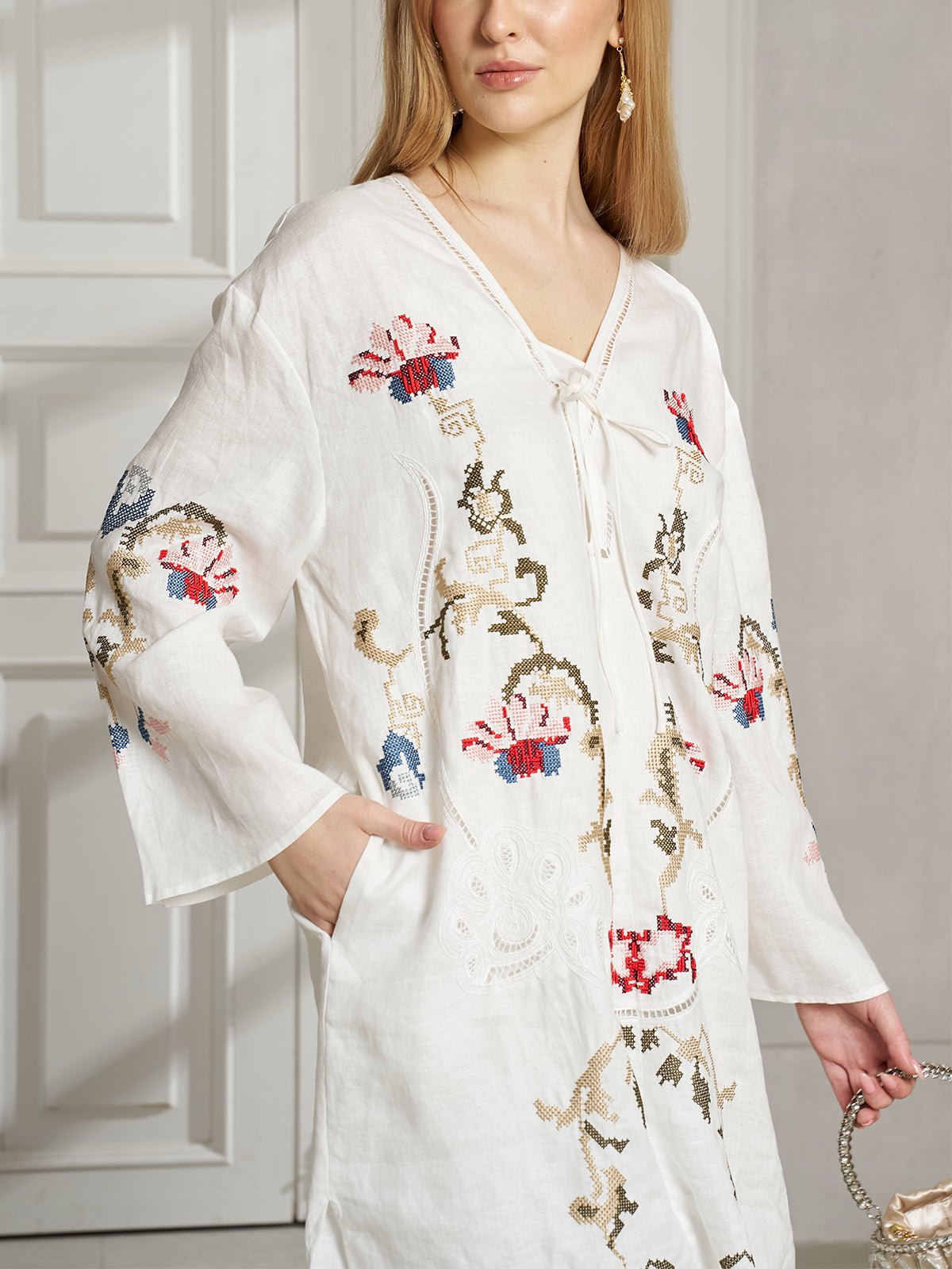 A woman stands indoors near a white door, holding a woven bag and wearing the Social Queen Botanical Embroidered Linen Top, featuring ethnic-inspired floral and abstract embroidery for an effortless resort style look.