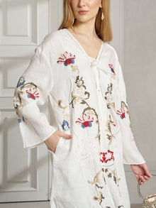 A woman stands indoors near a white door, holding a woven bag and wearing the Social Queen Botanical Embroidered Linen Top, featuring ethnic-inspired floral and abstract embroidery for an effortless resort style look.