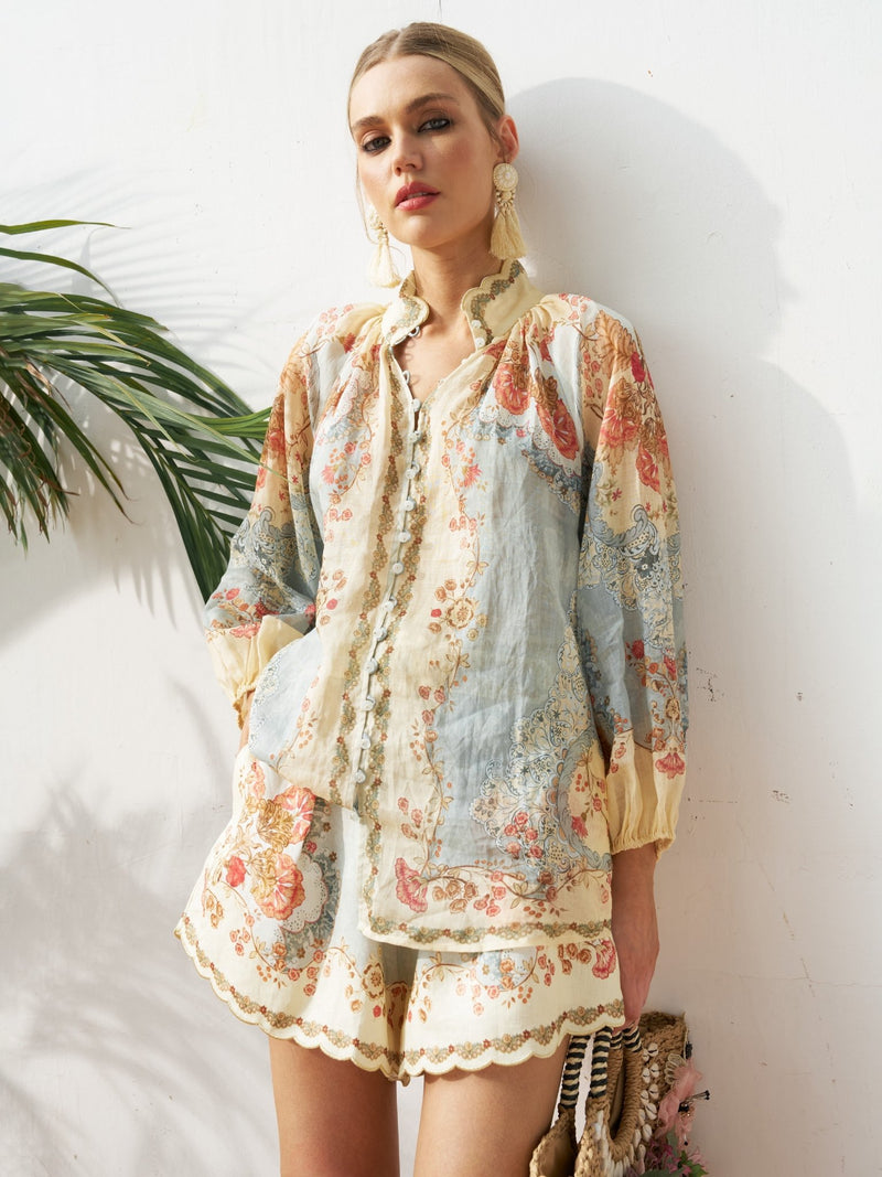 A woman wears the Social Queen Botanical Placement Print Ramie Blouse, featuring a light floral design and lantern sleeves, paired with matching shorts, standing against a white wall with a green palm leaf behind her.