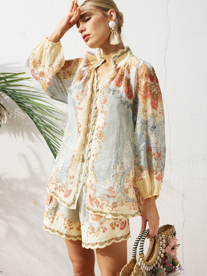 A woman stands against a white wall in the Social Queen Botanical Placement Print Ramie Blouse with lantern sleeves and shorts, holding a straw bag and shielding her face from the sun.