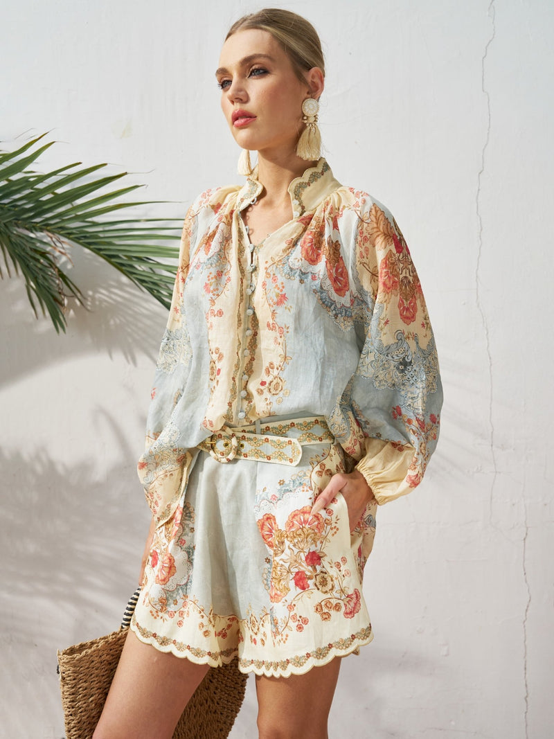 A woman stands against a white wall wearing Social Queen's Botanical Placement Print Ramie Shorts and a matching blouse, styled with statement earrings, a belt, and a woven handbag.