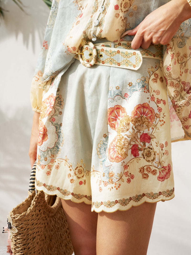 Wearing Social Queen’s Botanical Placement Print Ramie Shorts with a matching belt and blouse, holding a woven bag; the ramie fabric features scalloped edges and intricate floral patterns.
