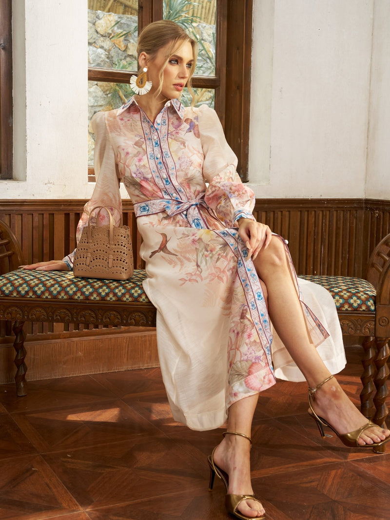 A woman in the Social Queen Botanical Placement Silk Shirt Dress sits on a wooden bench with a tan handbag, gold sandals, and statement earrings, one leg crossed over the other.