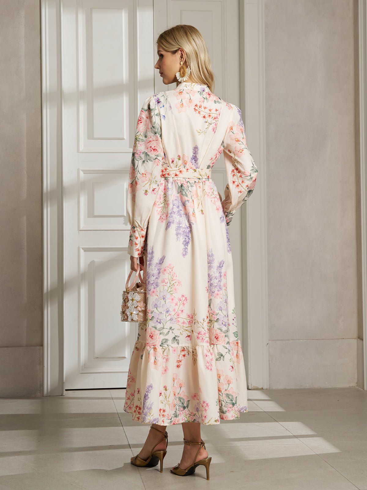 A woman indoors, facing away, wears the Social Queen Botanical Print Cotton-Linen Dress with long sleeves and heels, holding a small handbag.