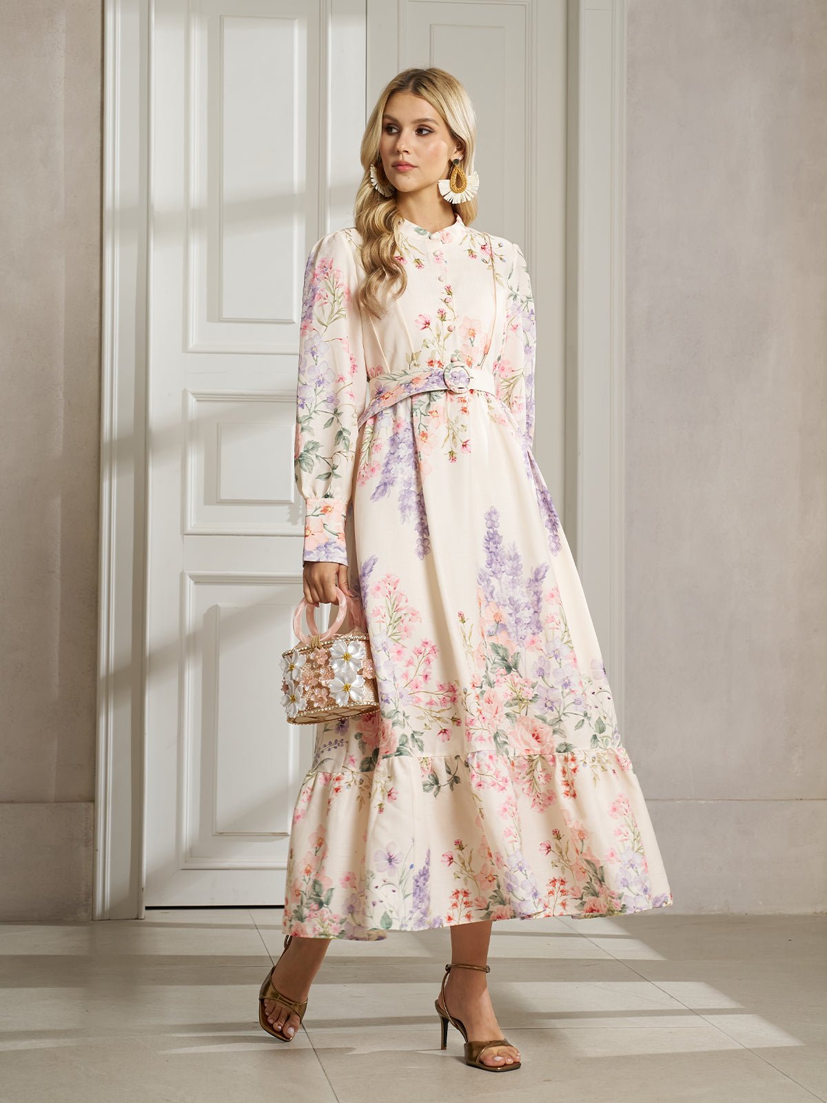 A woman stands indoors wearing the Social Queen Botanical Print Cotton-Linen Dress, featuring long sleeves, a belted waist, and a vintage-inspired maxi silhouette. She accessorizes with a small handbag and heeled sandals.