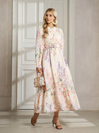 A woman stands indoors wearing the Social Queen Botanical Print Cotton-Linen Dress, featuring long sleeves, a belted waist, and a vintage-inspired maxi silhouette. She accessorizes with a small handbag and heeled sandals.