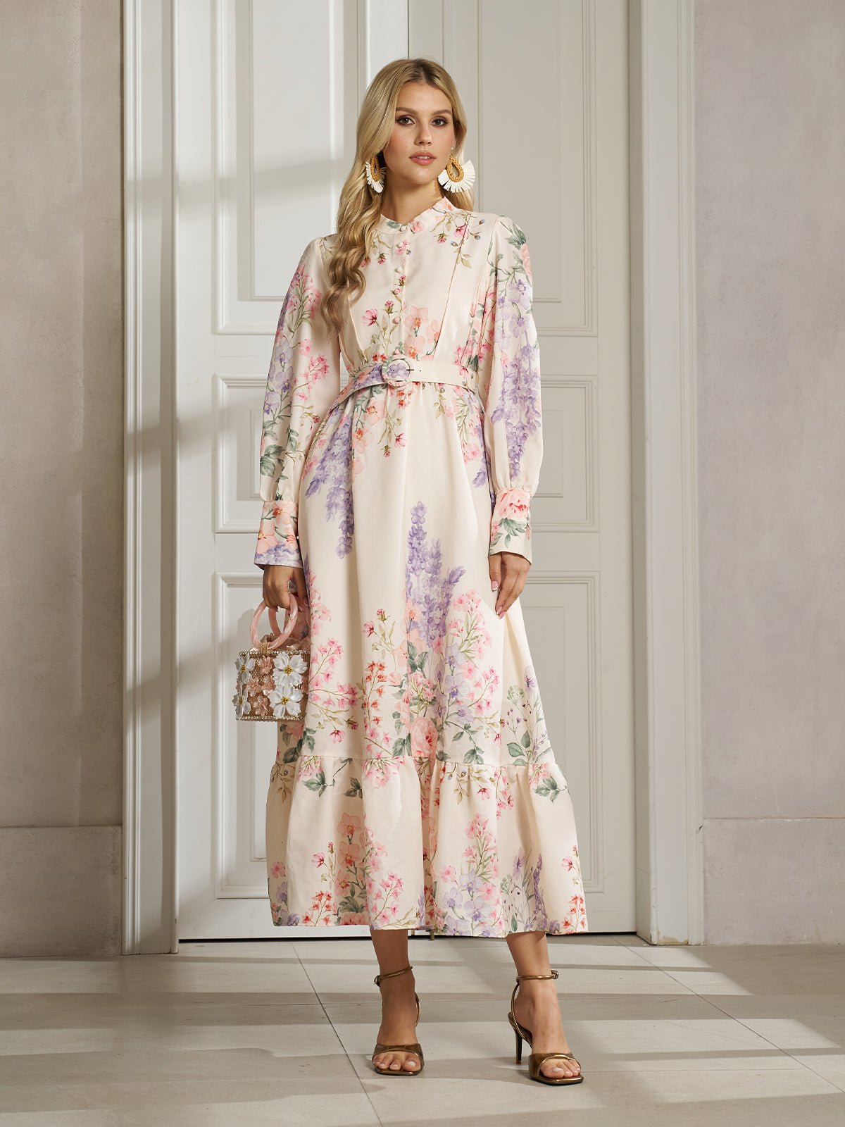 A woman with blonde hair wears the Social Queen Botanical Print Cotton-Linen Dress with a belt, paired with heels and statement earrings, standing indoors by a white door.