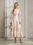 A woman with blonde hair wears the Social Queen Botanical Print Cotton-Linen Dress with a belt, paired with heels and statement earrings, standing indoors by a white door.