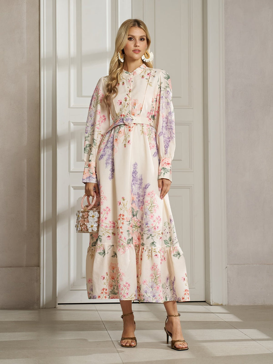 A woman with blonde hair wears the Social Queen Botanical Print Cotton-Linen Dress with a belt, paired with heels and statement earrings, standing indoors by a white door.