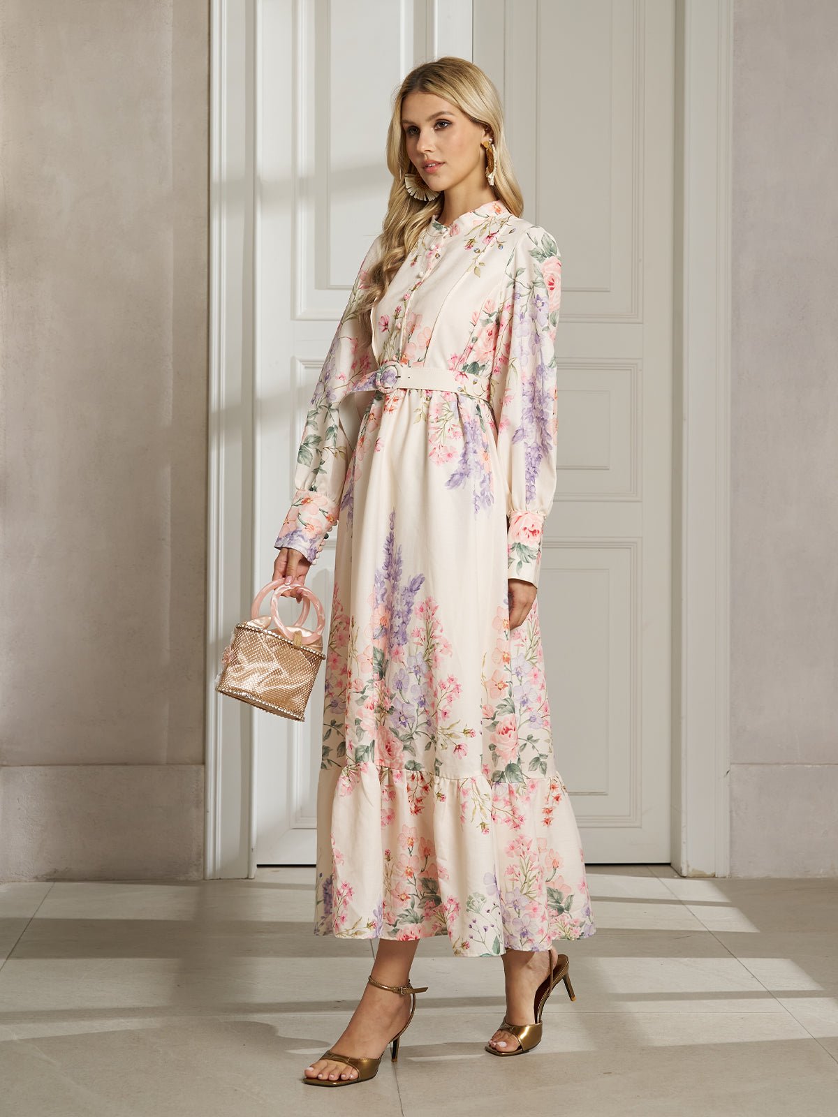 A woman in the Social Queen Botanical Print Cotton-Linen Dress with long sleeves, gold sandals, and a small woven handbag stands in an elegant room with white walls and doors.