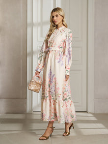 A woman in the Social Queen Botanical Print Cotton-Linen Dress with long sleeves, gold sandals, and a small woven handbag stands in an elegant room with white walls and doors.