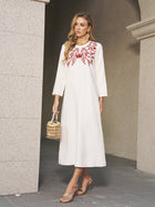A woman stands by a stone wall in the Social Queen Butterfly Embroidered A-Line Dress, styled with gold earrings, heels, and a gold handbag—a chic choice for any wedding guest.
