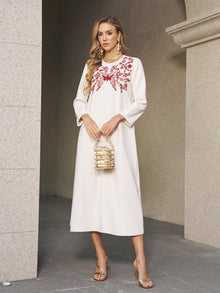 A woman in the Butterfly Embroidered A-Line Dress by Social Queen, paired with a gold handbag, stands against a stone wall—an ideal choice for any wedding guest.