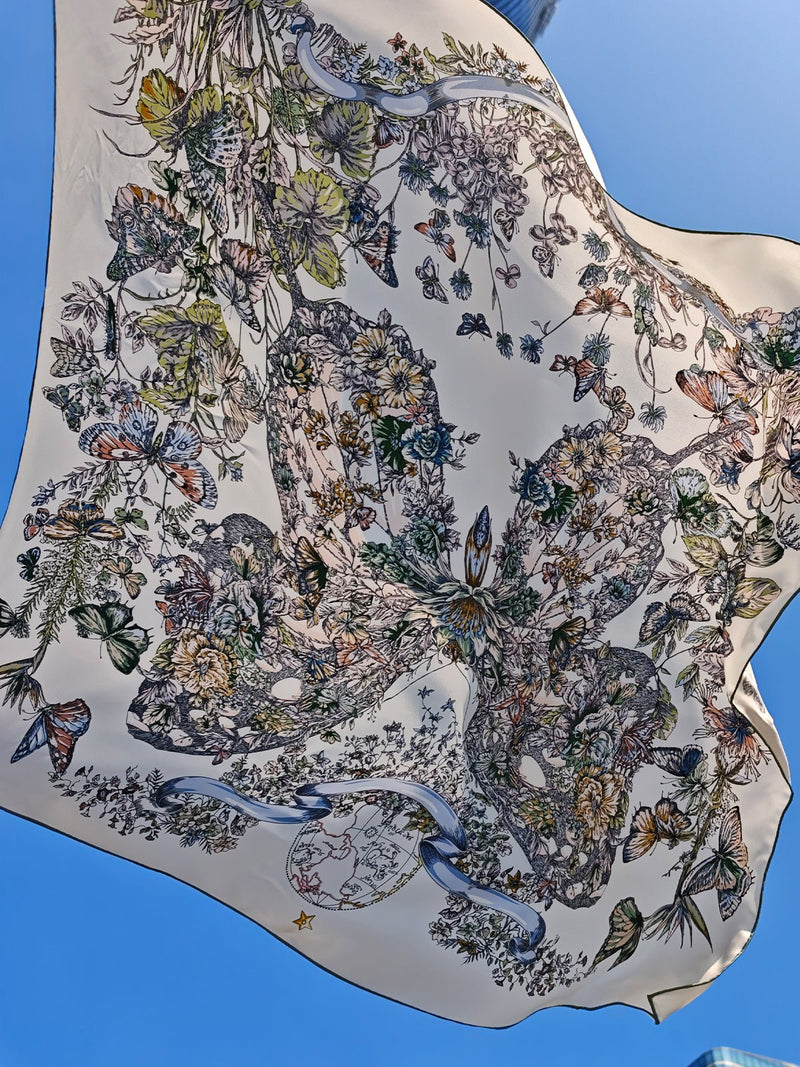 The Social Queen Butterfly Print Silk Scarf, crafted from mulberry silk and adorned with detailed floral and butterfly motifs, billows gracefully in the wind beneath a clear blue sky.