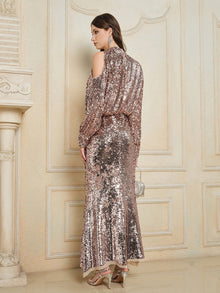 A woman stands indoors facing away, wearing the Social Queen Champagne Sequin Off-Shoulder Top, paired with silver high heels and holding a small clutch bag.