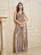 A woman stands against an ornate cream-colored wall, wearing Social Queen’s Champagne Sequin Wide-Leg Pants and holding a small beige clutch.