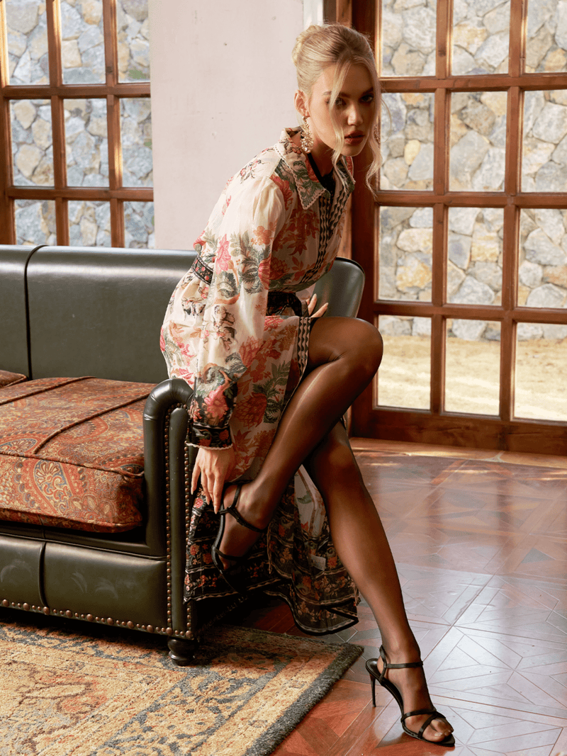A woman in a Social Queen Color-Block Floral Embroidery Dress and black heels sits on the edge of a green couch, adjusting her shoe, with large windows and stone walls in the background.