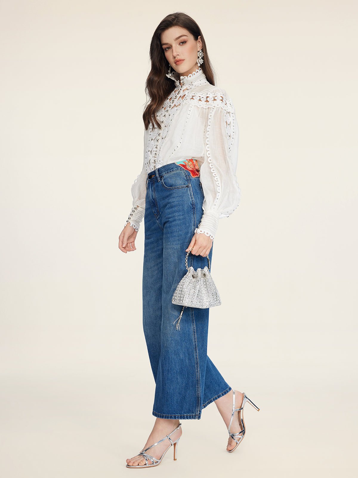 A woman in profile wears a white lace blouse, Social Queen Colorblock Floral Embroidery Jeans, silver heels, and holds a small silver handbag against a plain background.