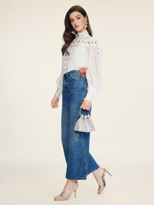 A woman in profile wears a white lace blouse, Social Queen Colorblock Floral Embroidery Jeans, silver heels, and holds a small silver handbag against a plain background.