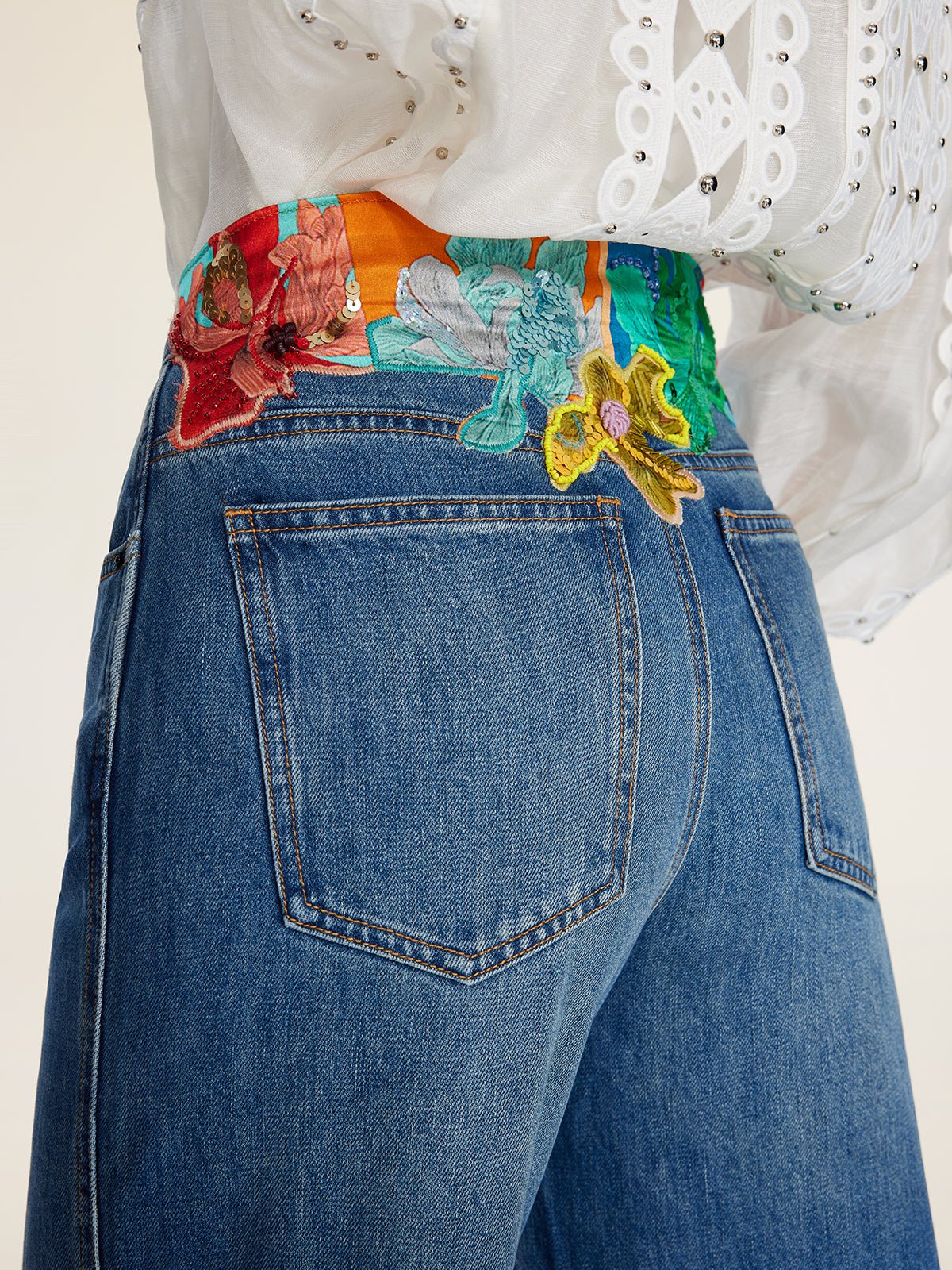 Close-up of a person wearing Social Queen Colorblock Floral Embroidery Jeans featuring high-waist, wide-leg, hand-distressed denim and floral embroidery at the waist, paired with a white eyelet blouse.