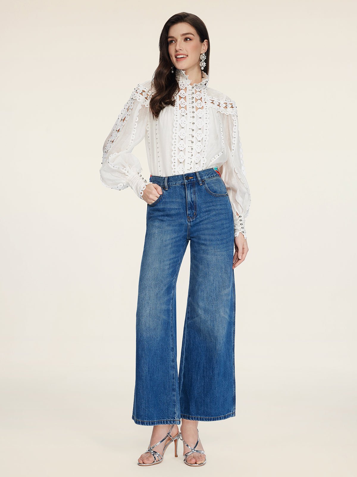 A woman in a white lace blouse, Social Queen Colorblock Floral Embroidery Jeans, and silver heeled sandals stands against a plain beige background.