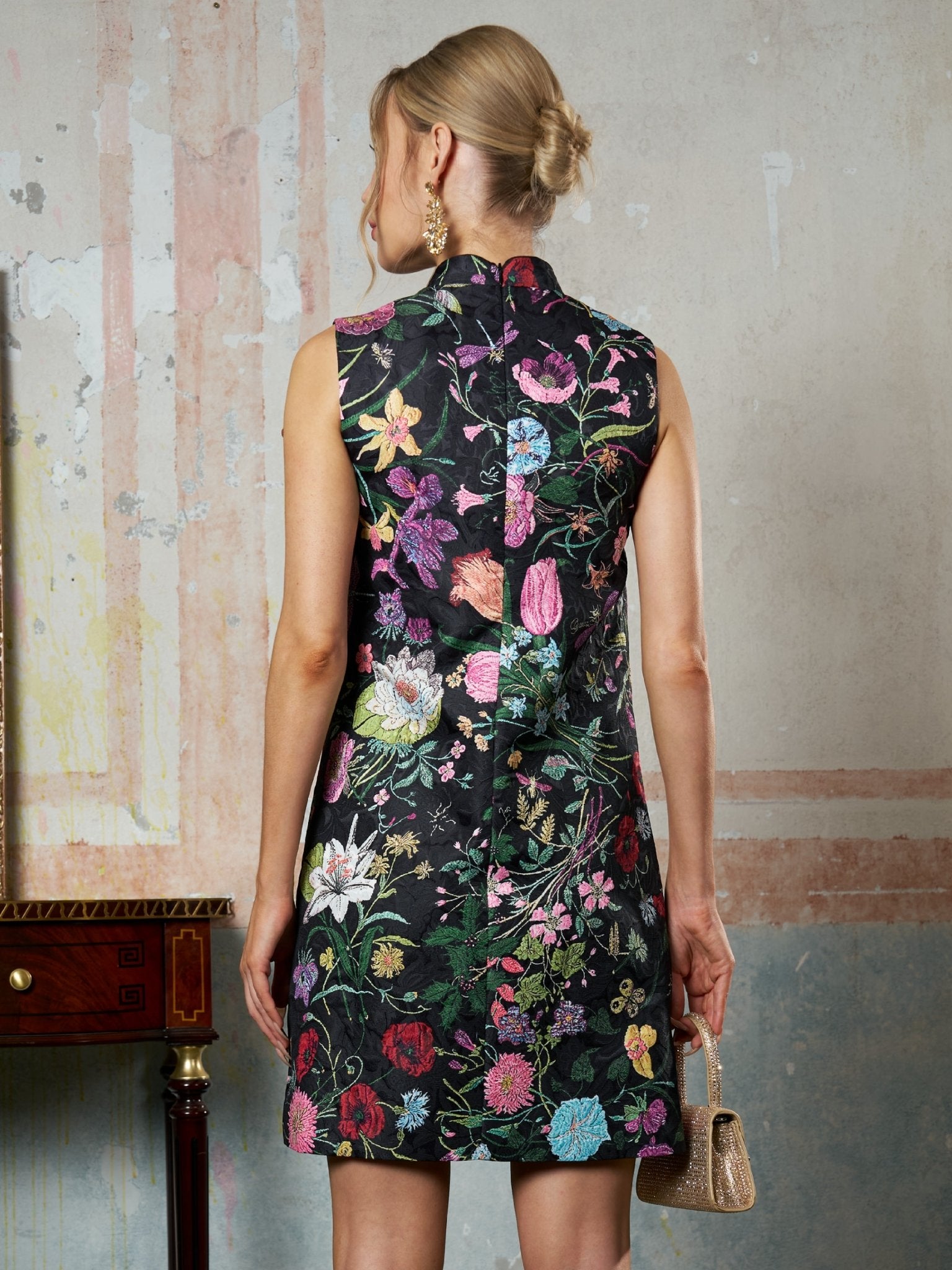 A woman faces away in Social Queen’s Colorful Floral Mandarin Collar Dress, holding a small textured handbag, standing by a vintage table against a faded, patterned wall.